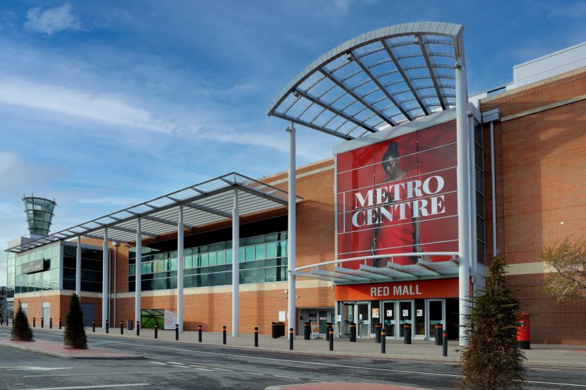 Exterior of Metrocentre. The large, boxy, brick structure has metal accents creates a frame for the entrances. Large red 'Metro Centre' sign above the entrance