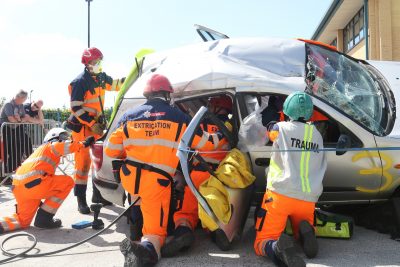 Vehicle on its side with smashed windscreen surrounded by trauma and extrication team wearing full orange high viz and helmets. The teams are working to get people out of the car (this is a simulation as part of the UKRO challenge)