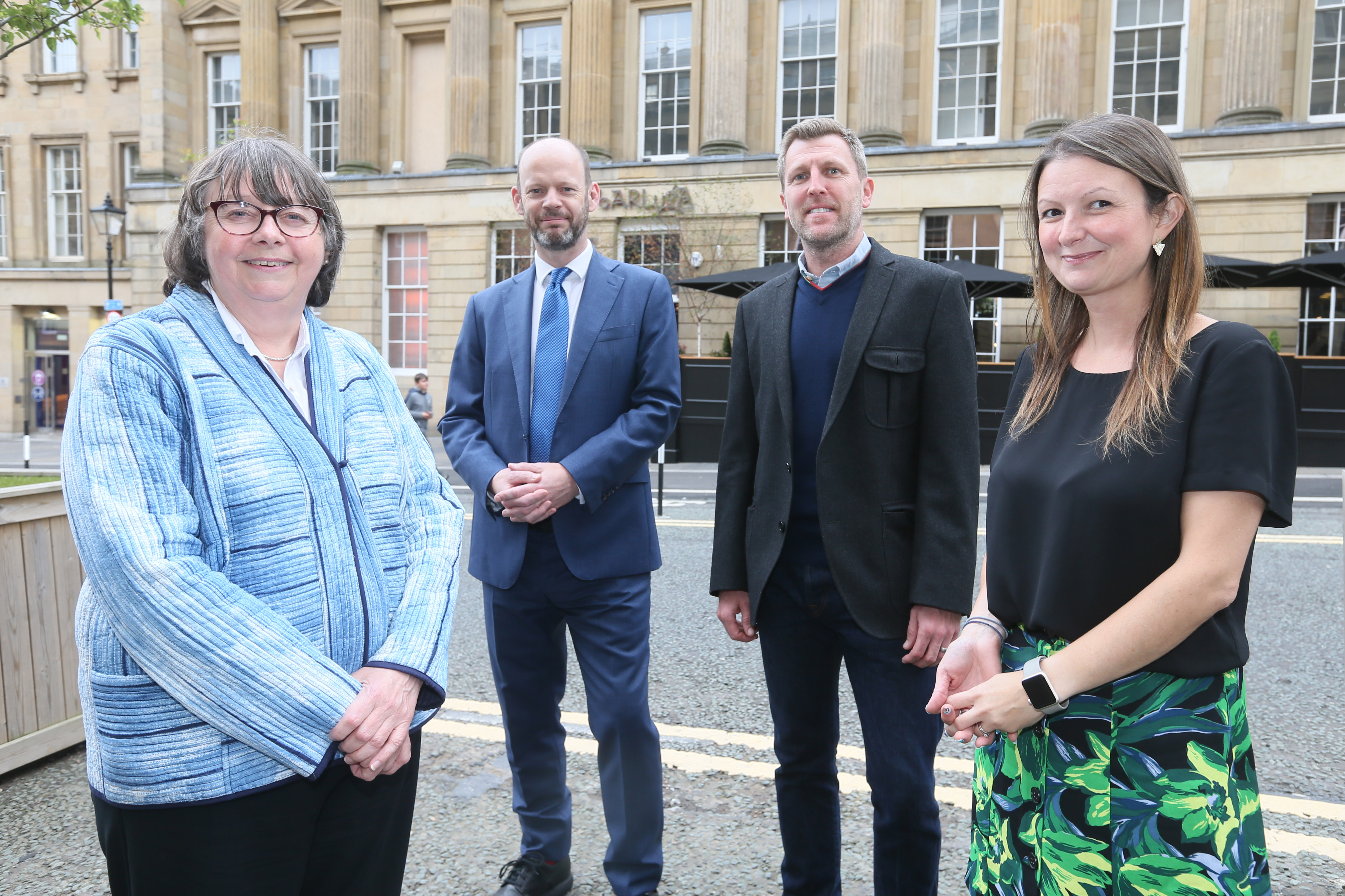 Four people stood smiling towards camera with sandstone building in background. On the left stands a woman in a light blue blazer with a short bob haircut and glasses. Next to her stands a man with no hair in a blue suit and tie and white shirt, he has a beard. Next to him stands a man with a grey blazer and blue jumper with light, cropped hair. Next to him stands a woman with longer brown hair, a black blouse and green tropical print skirt. They are all smiling towards the camera.