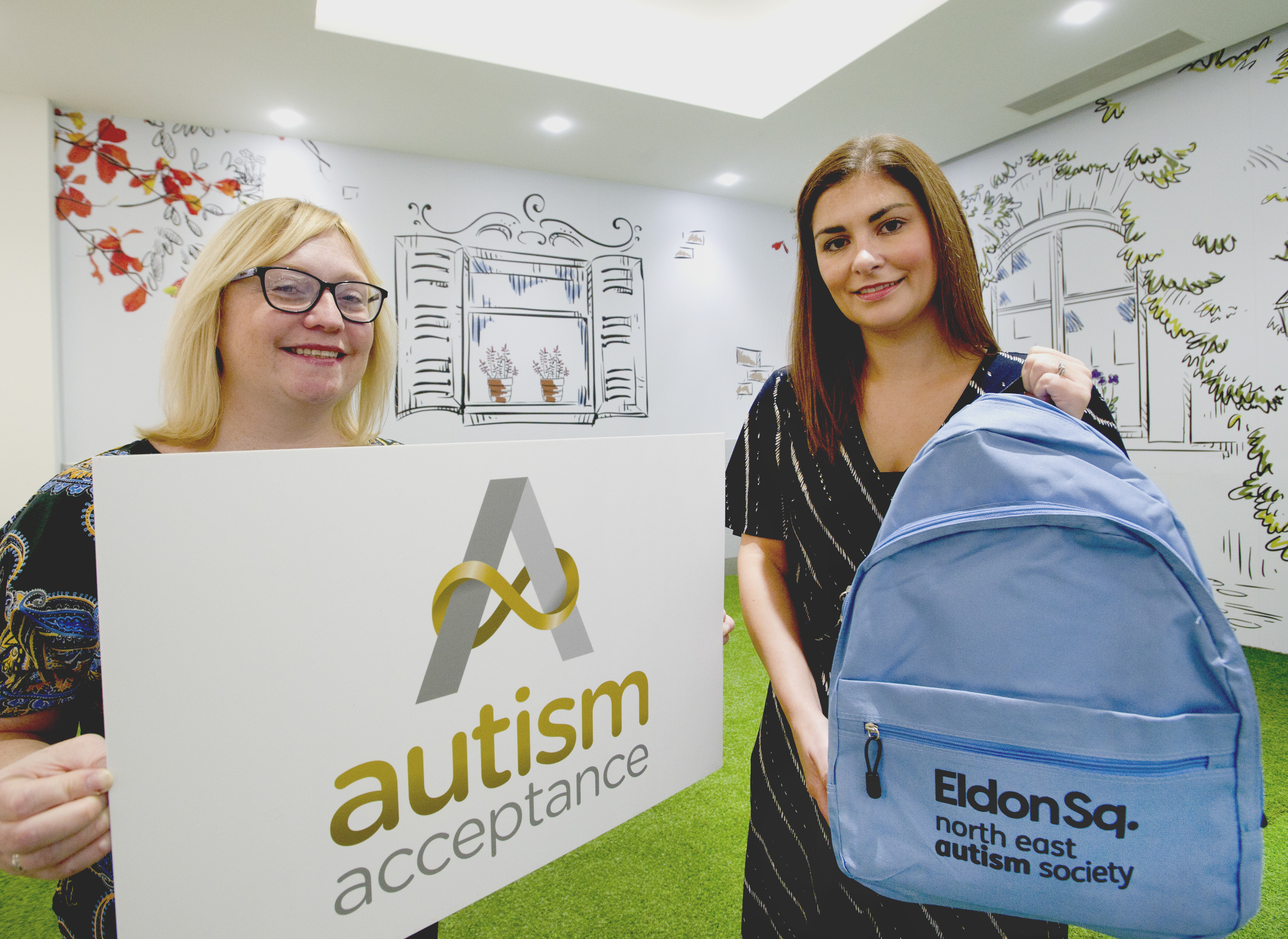 Two women stand smiling at camera. On the left a woman holds a large sign reading 'autism acceptance' and on the right the woman holds a light blue backpack which reads 'Eldon Sq. North East Autism Society'