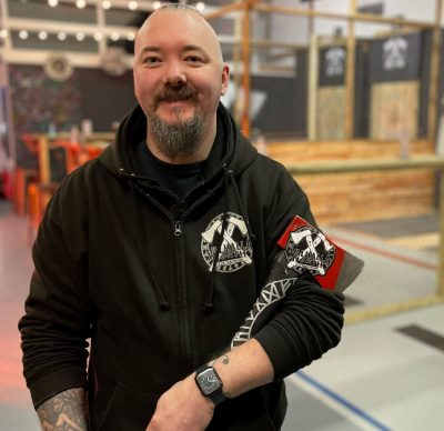 Man in black hoodie smiles to camera with hatchet in arm