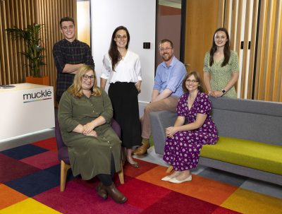 Six members of Muckle team including new appointment Alison in their office smiling to the camera. Three people are sat and three are stood.