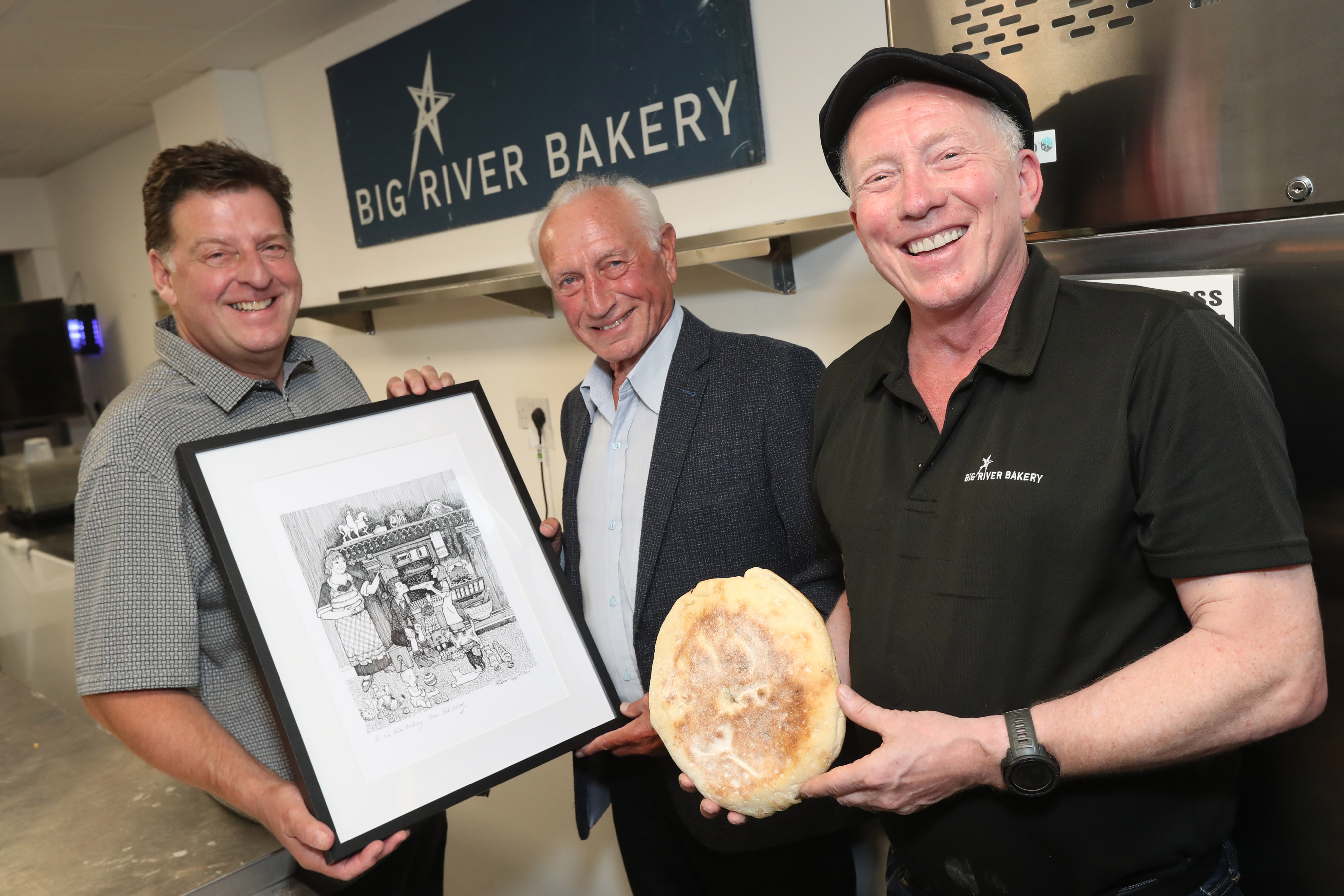 Three people stood inside Big River Bakery. Two are holding framed artwork and one is holding a stottie and smiling towards camera.