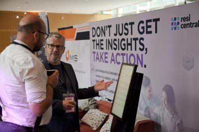 Two men stood at an exhibition stand talking next to a screen. One holds a coffee cup.