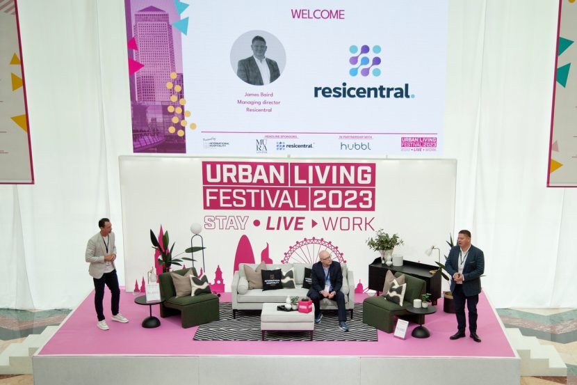 Three people on a pink stage in an airy room presenting at Urban Living Festival 2023. One is sat on the sofa in the middle and two are stood either side of the stage.