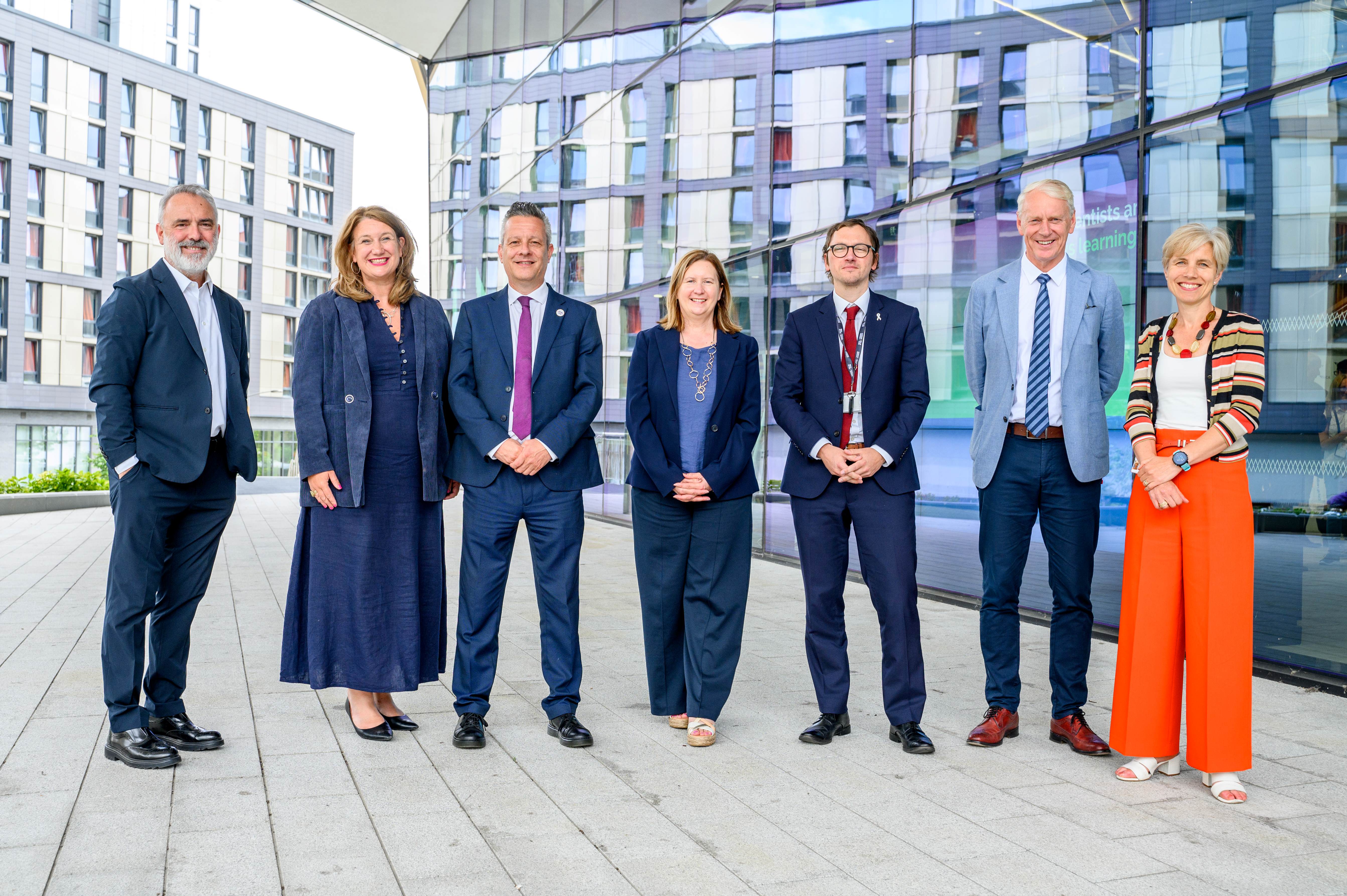 Seven of Newcastle's public and private sector city leaders stood in a row smiling at camera in front of glass building in suits and dresses.