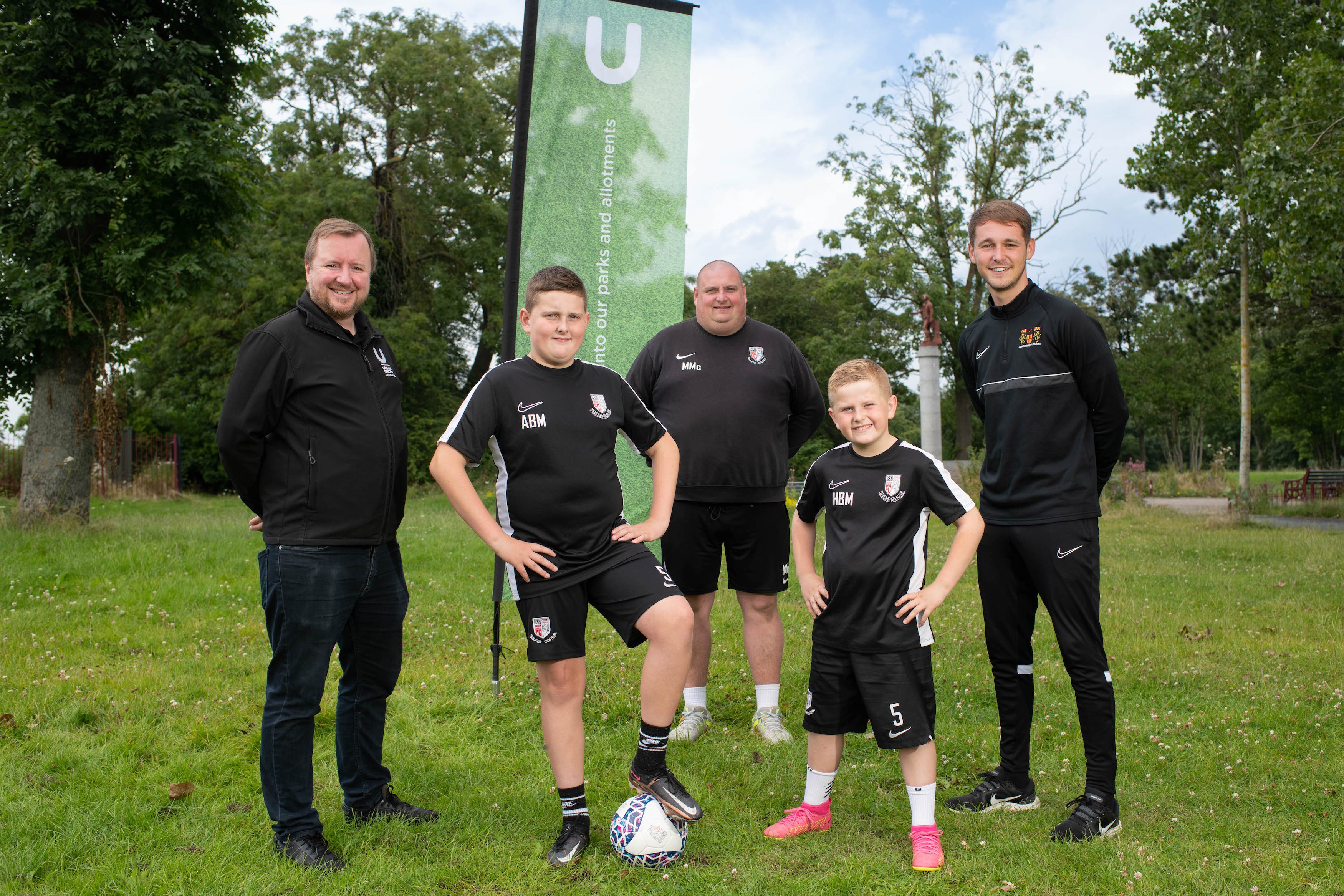 Group of adults and two kids stood in grassy. leafy park with football and sports kit on.