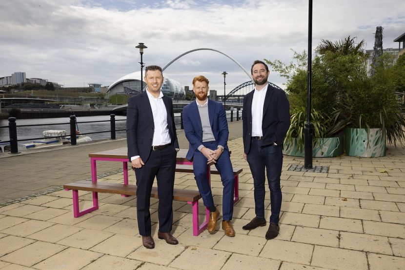 L-R, Jamie, Sam and Matthew, at the Quayside, Newcastle. They are smiling at the camera with the river Tyne and the Millennium Bridge in the background.