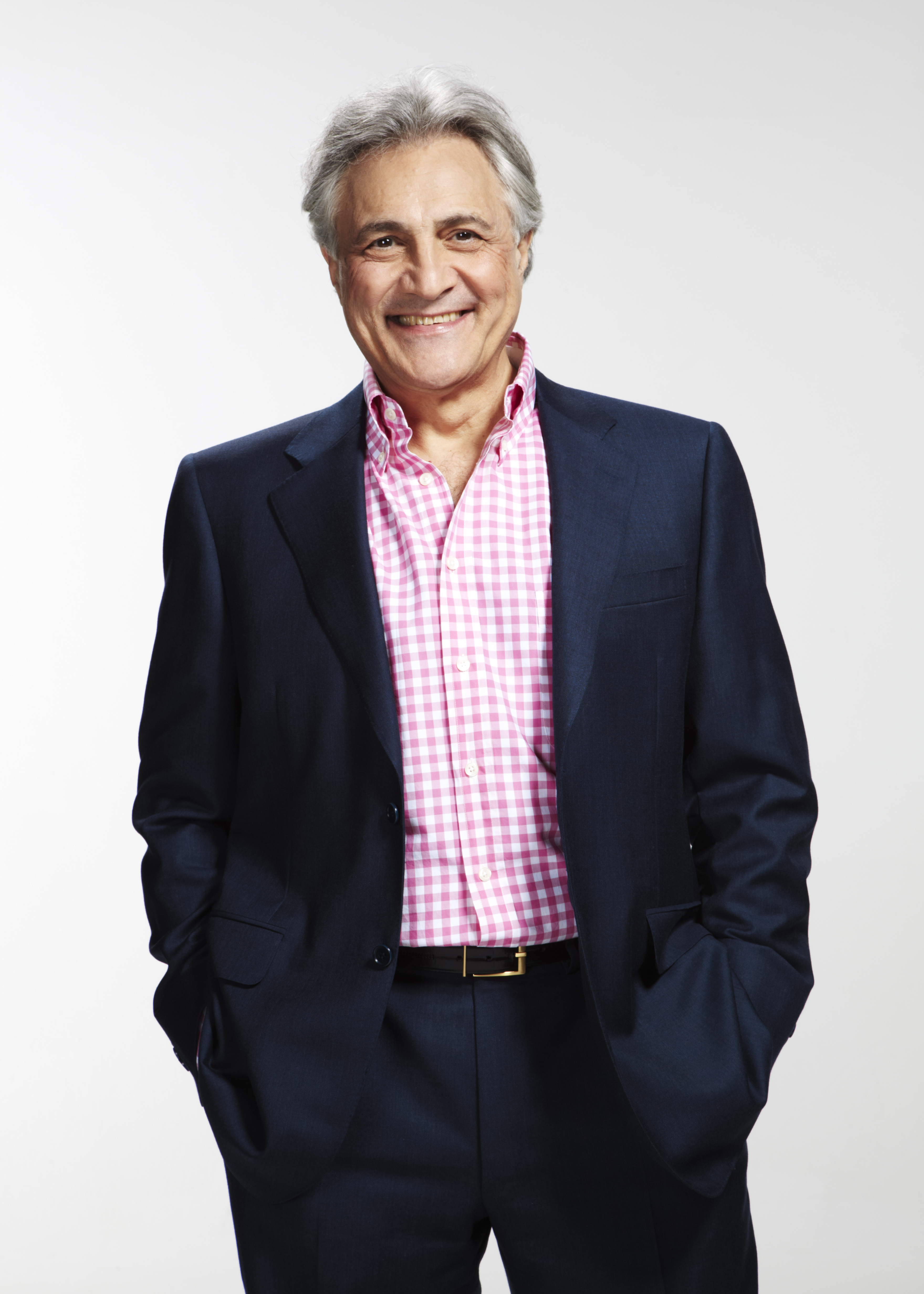 John Suchet standing with hands in his pockets smiling at the camera.