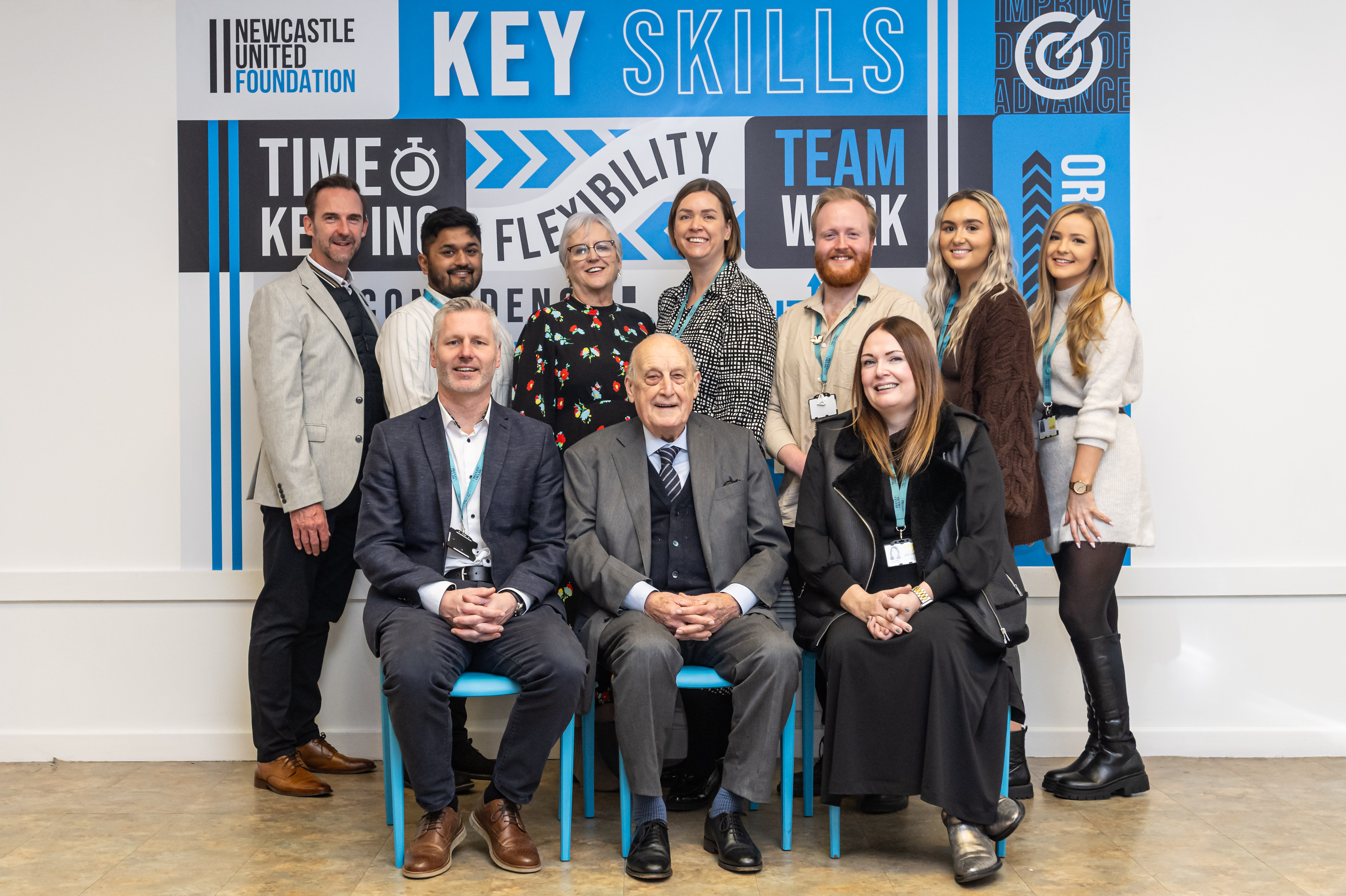 Two rows of people in front of Newcastle United Foundation banner smiling towards camera.