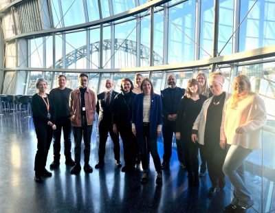 Group of people stood inside The Glasshouse ICM with the Tyne Bridge in the background outside.