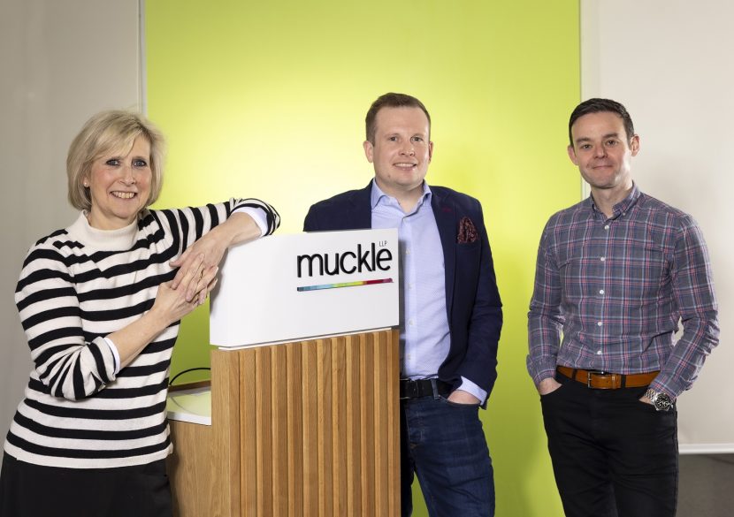 Three people stood around a Muckle LLP podium smiling towards the camera