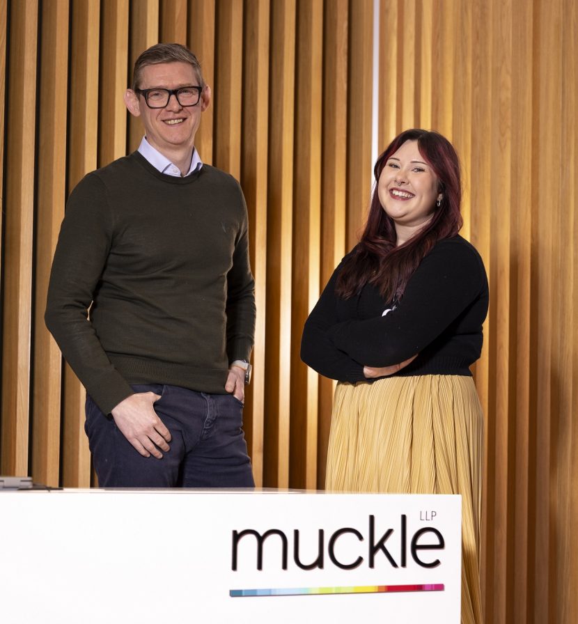 Anthony and Melissa stood in front of desk which has Muckle LLP logo along the top.