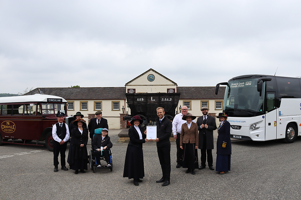 Beamish staff in costume stood outside of the museum entrance being presented the award by CPT representative with coach on the right and older bus on the left.