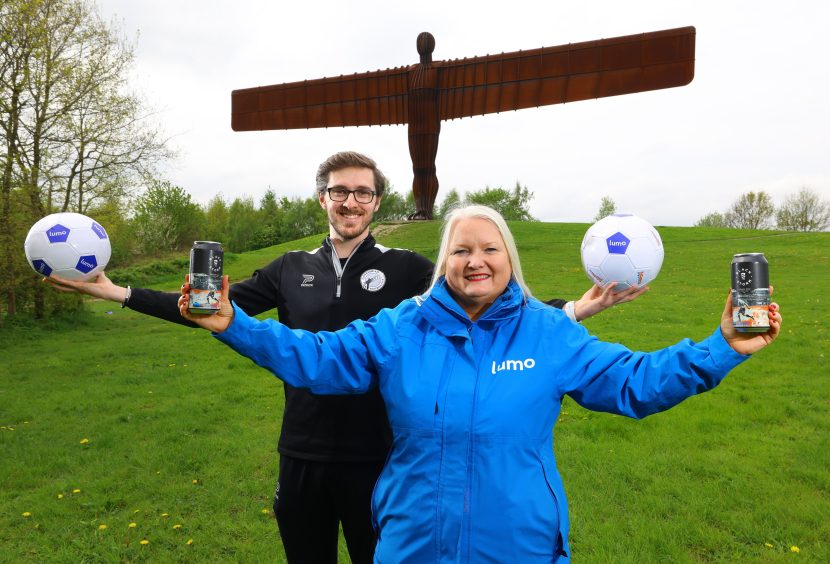 Two people stood in front of Angel of the North. Both have their arms outstretched mimicking the statue and one person is holding a football in each hand and one is holding a can of beer in each hand.