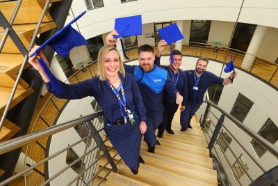 Four Lumo employees stood on some stairs holding blue graduation caps and smiling to camera.