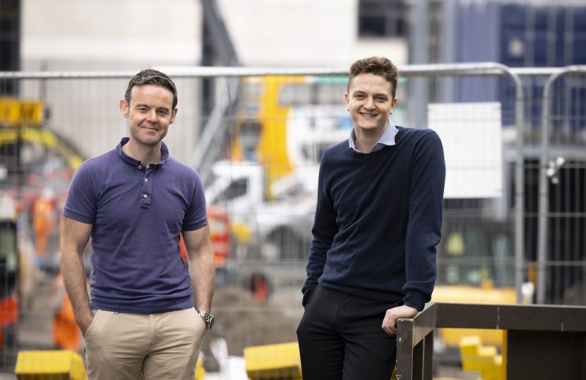 Photo of Adam and Kieran stood near a construction site smiling to camera