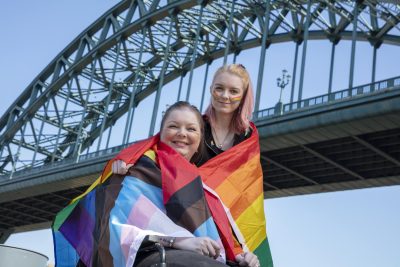 Two people wrapped in Pride flags hugging one another and smiling to camera with the Tyne Bridge in the background.