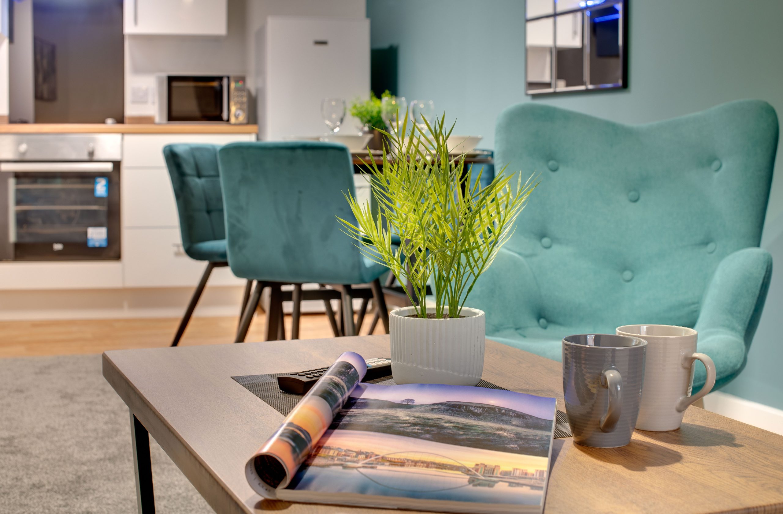 Interior of serviced apartment with blue arms chair and coffee table in the foreground and kitchen and dining area in the background.