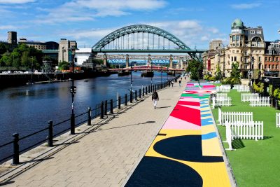 Long shot down the Newcastle Quayside with the river on the left and path in centre and brightly coloured mural down the centre.