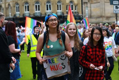 Group of young people walking along street in Newcastle in Pride Parade. One is holding a sign which reads: Free hugs
