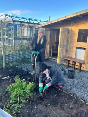 Two BeanHouse Events team members in a garden one with a standing with a spade in hand and another crouched and digging into the soil. Both are smiling to camera.