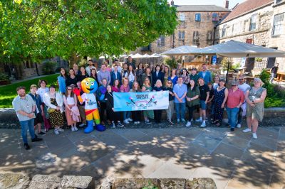 Large group gathered together smiling to camera in a restaurant garden with a Northern Pride banner and mascot.