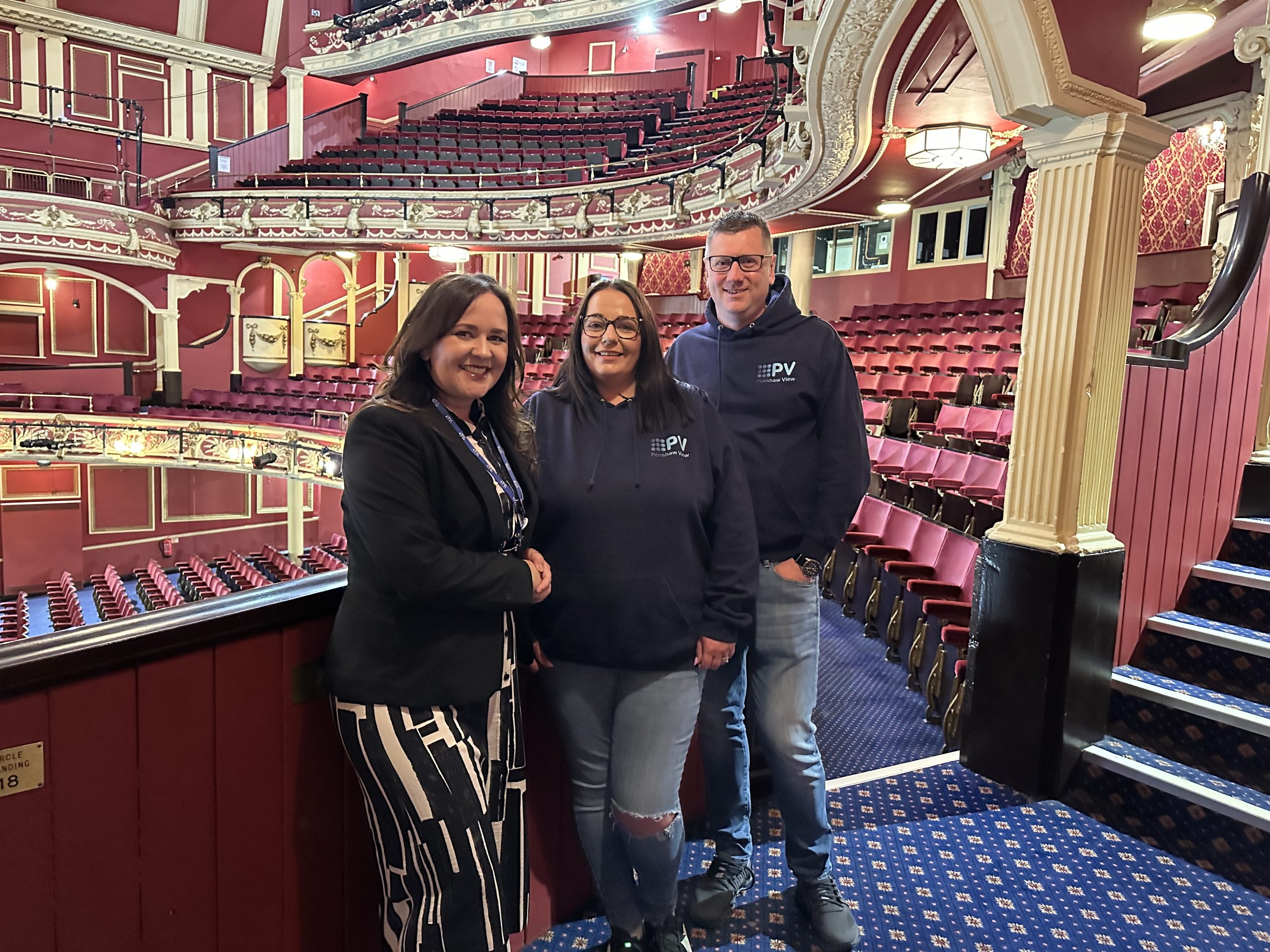 Three people stood smiling to camera inside the Sunderland Empire theatre which has traditional red seats and royal blue carpets.