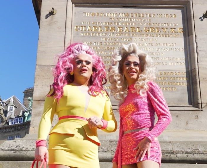 Two drag queens posing in front of Grey's Monument in brightly coloured wigs and dresses