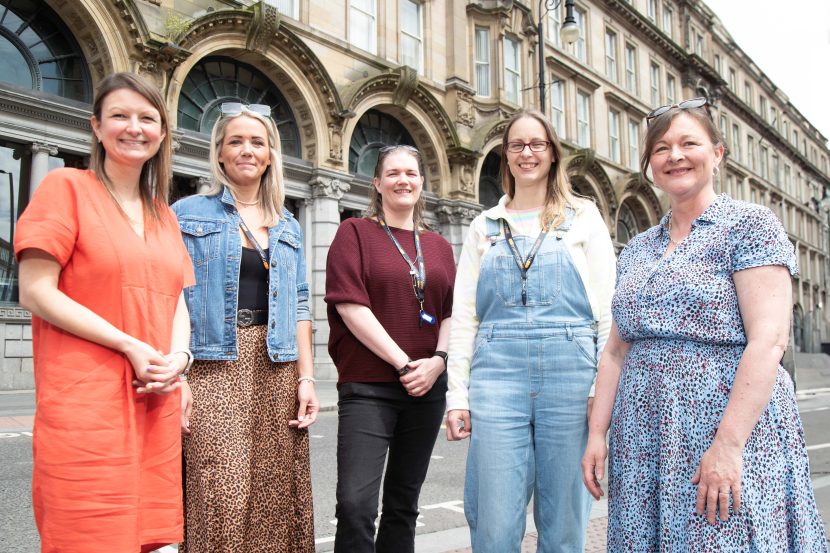 A photograph of five people smiling towards the camera. In the background are the Collingwood Buildings in Newcastle city centre.