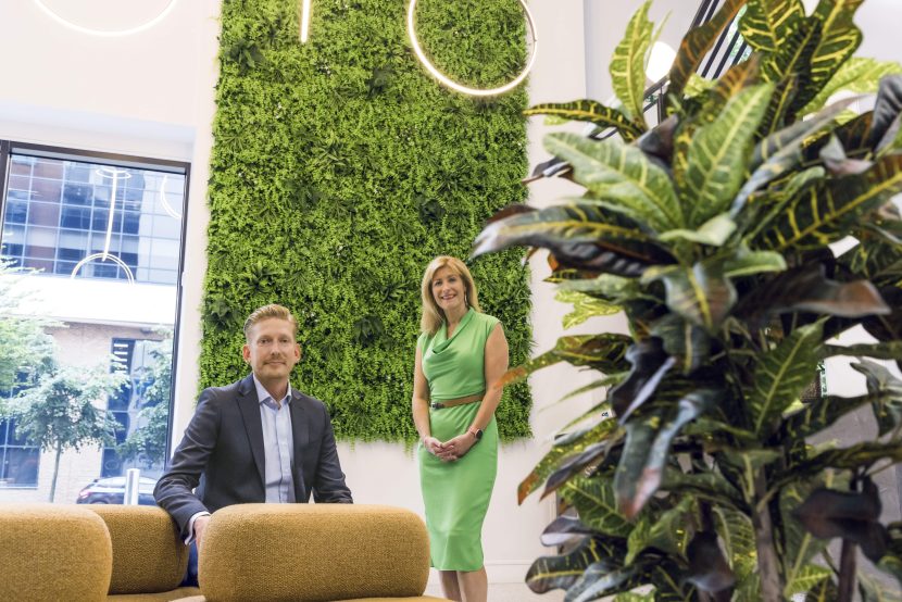 Alex and Andi posing in office building reception with living wall behind them and foliage to the right.