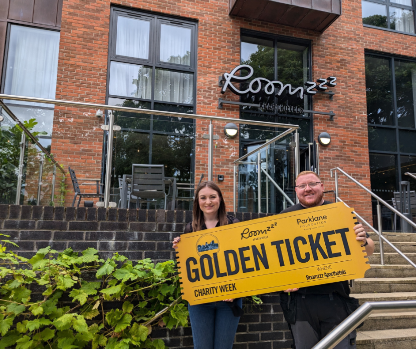 Two people holding large' Golden Ticket' outside of Roomzzz Aparthotel