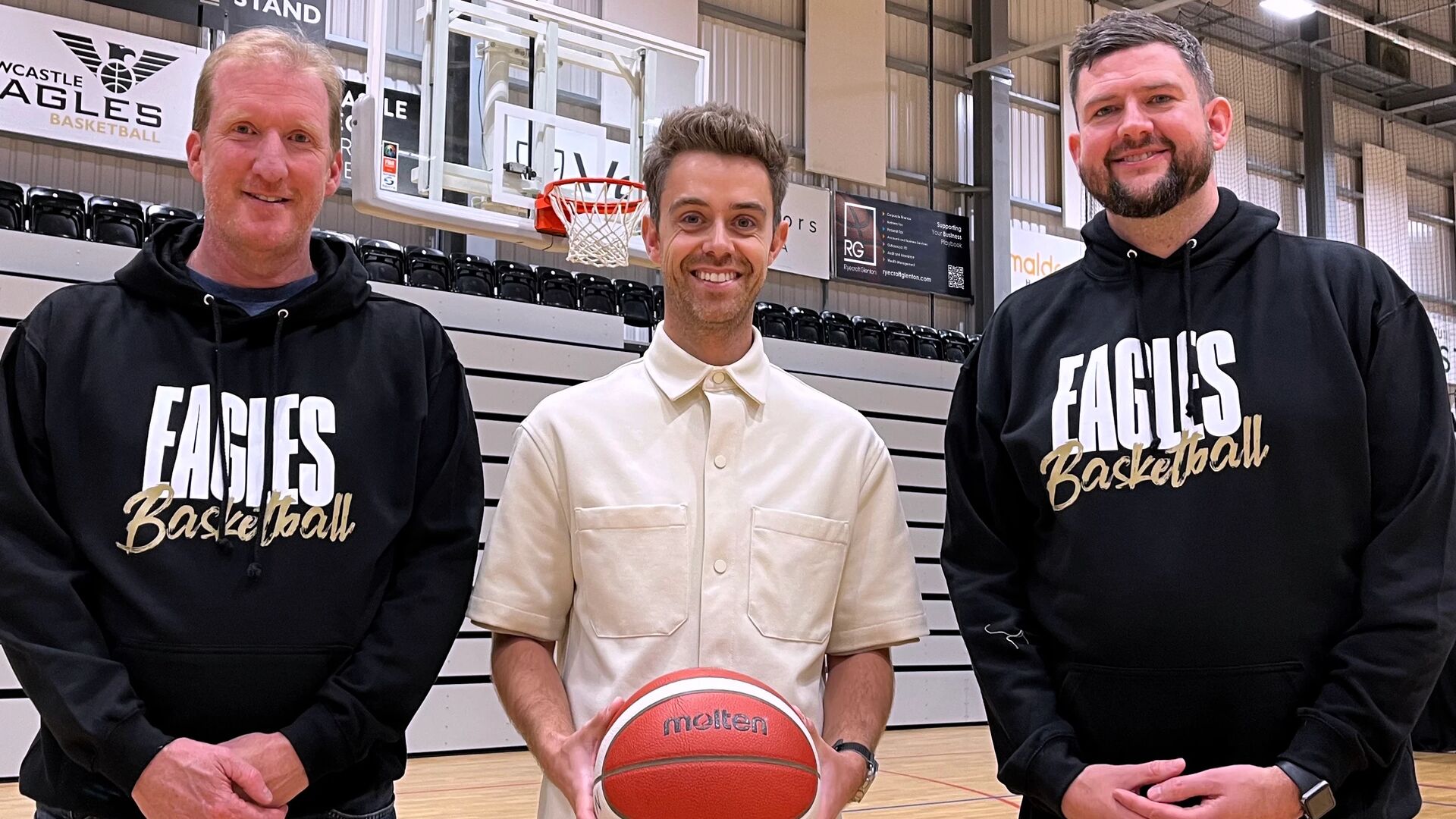 Three people stood on basketball court, person in the middle is holding a basketball and two on either side are wearing Newcastle Eagles hoodies.