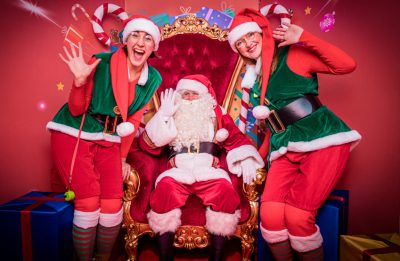 Person dressed as Santa sat on chair with two people dressed as elves stood on either side.