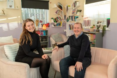 Lydia and Chris sat in a room filled with books and sat on light pink sofa and chair.